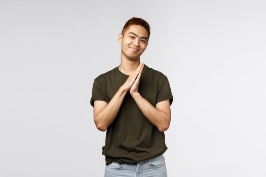 Portrait of cheerful, lovely asian man in greey t-shirt, show namaste gesture, asking for favour or appreciate help, clasp hands together and smiling, praying over grey background.