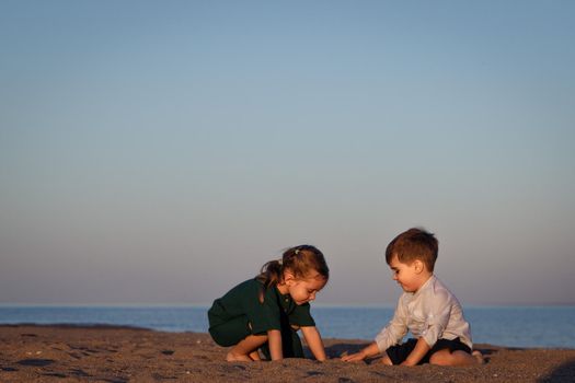 A girl and a boy 3-4 years old play with sand on the beach in the sunset light. Copy space.