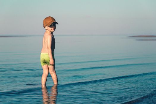 Boy in green shorts and brown baseball cap stands in the sea. Copy space. Toned minimalism photo.