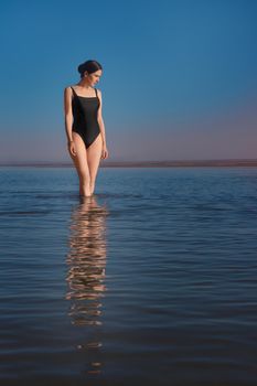 Young beautiful slim woman wearing a black swimsuit stands in the sea and watching sunset. Copy space vertical