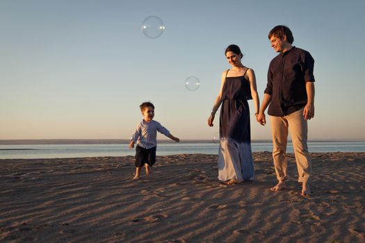A young family with a child walks along the beach in the evening natural light, soap bubbles fly around.