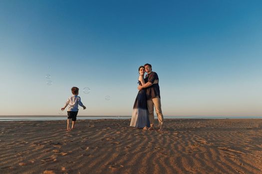 Young couple with their little son are walking along the beach in the evening natural light, soap bubbles fly around. Copy space.