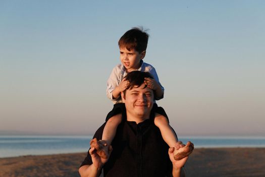 Portrait of a cute boy 3 years old on his father's neck walking on the beach in sunset lighting.