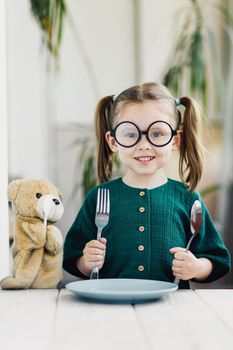 Little beautiful girl in green muslin dress waiting breakfast with bear toy. Little cute girl at white dining table in kitchen. Healthy nutrition for young kids.
