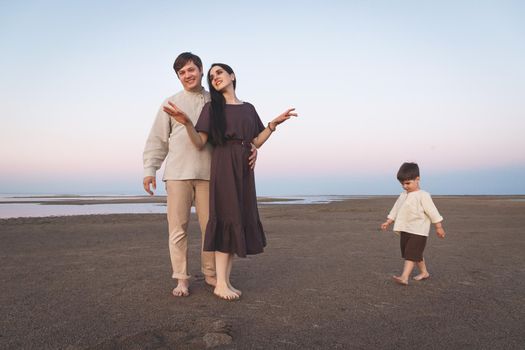 Young family with a three year old son walks barefoot along the wild evening beach. Family look linen clothes.