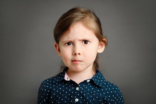 Perplexed little caucasian kid girl 4 -6 years old in a blue dress with polka dots on grey background studio portrait. Bewilderment emotion cute girl headshot