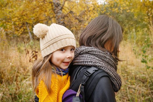 Young mother with her toddler kid girl on back in ergonomic baby carrier in autumn nature. Babywearing and active mother concept.