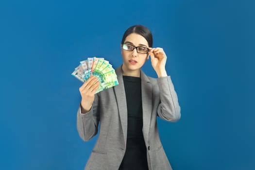 Young woman in glasses looks at a pack of tenge with interest on blue background.