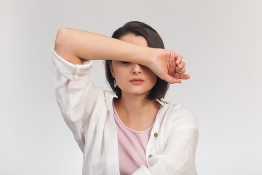 High-key studio portrait of a young woman in light clothes covering her eyes with her hand.