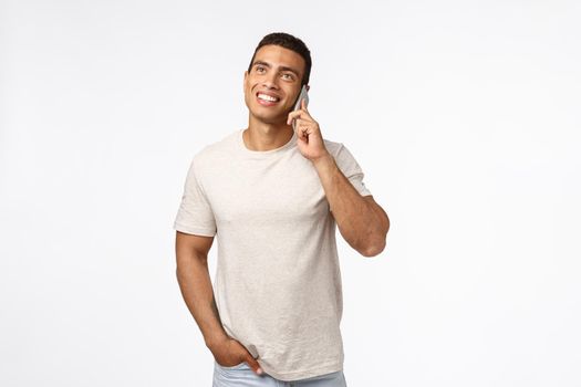 Cheerful, smiling young man in t-shirt, having conversation with friend, looking up enthusiastic, hold hand in pocket casually, smartphone pressed to ear, talking joyfully, white background.