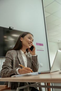 Smiling business asian woman working in office talking phone and making notes. High quality photo