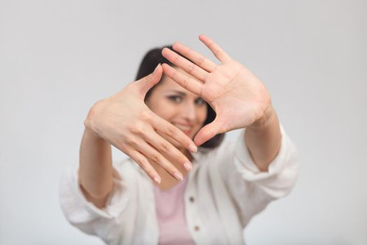 High key portrait of a young smiling woman looking at the camera through her hand framing. Selective focus on hands.