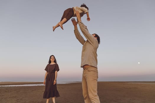 Young parents playing with their child on the beach by the sea. Father fun throws up son in the air.