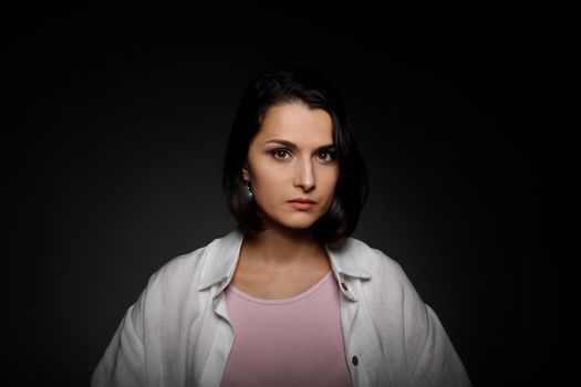 Low key portrait of serious young brunette multiethnic woman in pink tank and white shirt looking at camera.