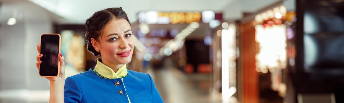 Cheerful woman flight attendant in aviation air hostess uniform looking at camera and smiling while holding smartphone