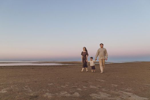 A young family with a little son in linen clothes are quietly walking along the evening beach. Copy space.