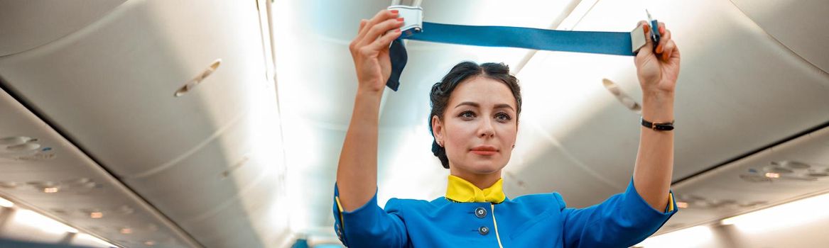 Woman stewardess in air hostess uniform holding safety belt while standing near passenger seats in airplane salon