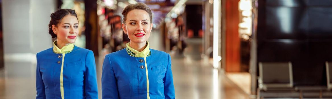 Two smiling women stewardesses in air hostess uniform walking down airport terminal