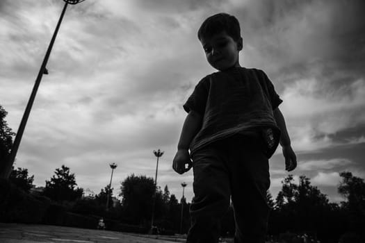 Black and white portrait of 3 year old boy in the park playing alone against the backdrop of a dramatic sky. BW lifestyle street photo.