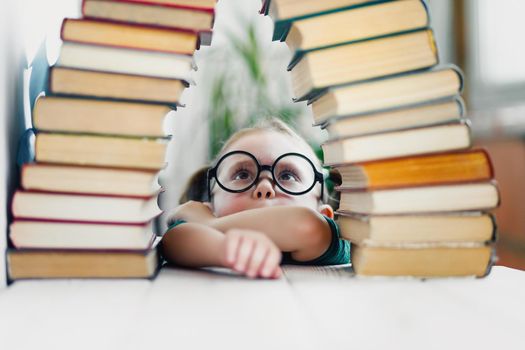 Beautiful funny little girl in round shaped glasses look up on many books, getting ready for school. Elementary education concept.