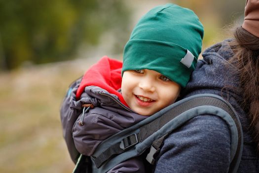 A smiling boy travels behind his mother in a baby carrier.