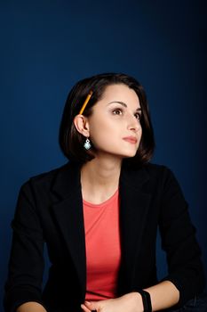 Vertical portrait of a young woman with a pencil behind her ear ponders new ideas on blue background.