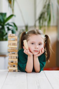 Portrait of beautiful sweet little girl in green dress with ponytails on her head holding a wooden block and wooden tower standing on a table.