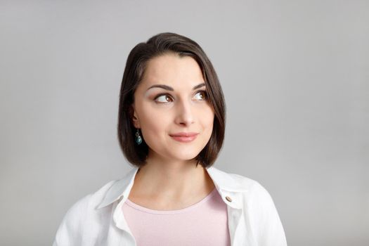 High key portrait of smiling young brunette multiethnic woman in pink tank and white shirt looking at top corner.