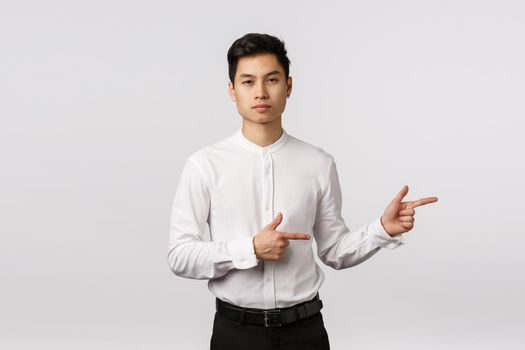 Serious-looking sassy and handsome asian millennial guy in formal wear, shirt and pants, pointing right, looking camera assertive, employer give direction, offer product, white background.