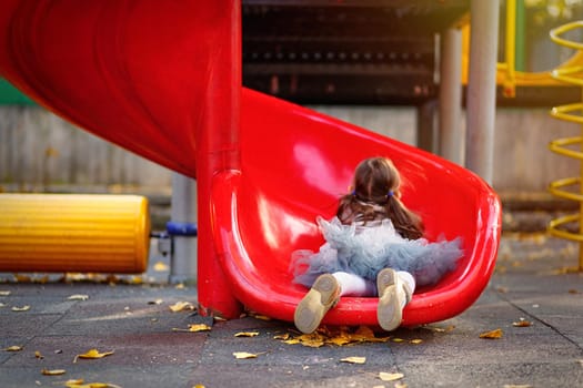 A cute girl in skirt tutu is riding a playground slide. Lifestyle outdoor portrait of a child. Motion blur and selective soft focus.