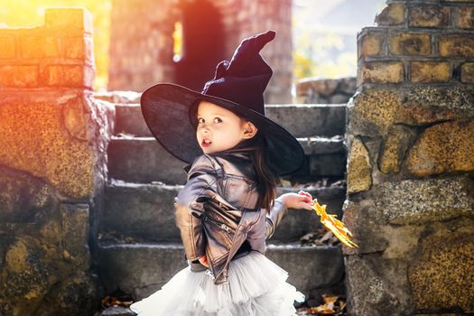 Emotional little girl with a yellow leaf in hand, in a witch hat walks through the stone castle, looks at the camera. Autumn and halloween concept