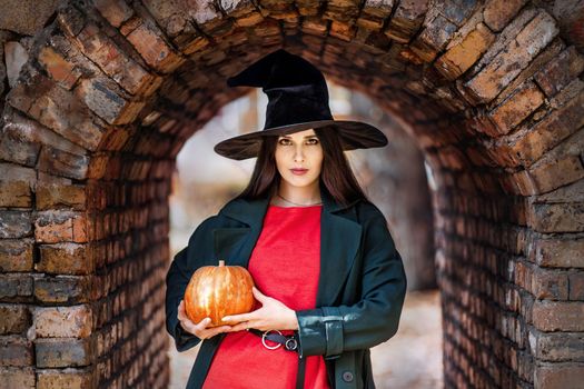 Pretty young woman outdoor portrait in witch hat holding a orange pumpkin. Halloween mood.