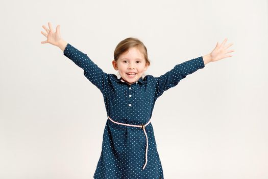 Happy little girl 4-6 years old wearing a blue dress with polka dots raised her hands in different directions, standing on a white background. Freedom, joy, school holidays