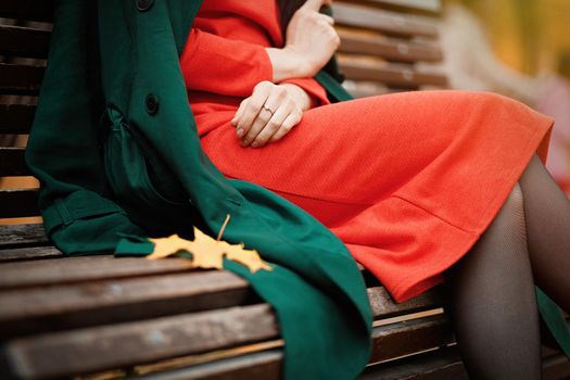 Woman in orange dress and green coat sitting on park bench outdoor. Autumn fashion.