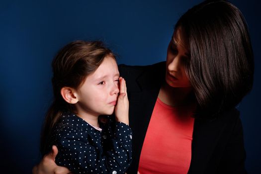 Young mother holding her crying kid girl on dark blue background.