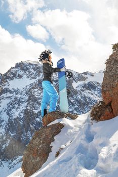 Freeride snowboarder woman in mohawk hat on top of rock in snowy mountain. Copy space, vertical