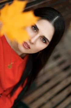 Portrait of a young brunette woman in an orange dress with a yellow autumn leaf out of focus. Autumn tones and mood vertical