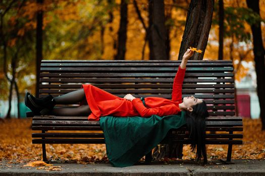 A young woman in an orange dress and a green coat lies on a bench in an autumn park and enjoys the autumn mood.