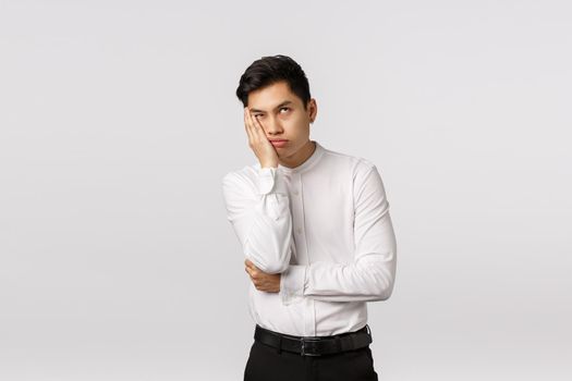 Bored and annoyed young asian businessman dying from boredom and annoyance, roll eyes up, facepalm, lean face on hand and waiting when meeting over, standing irritated white background.