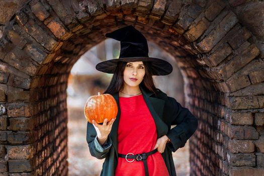 Beautiful young woman portrait in witch hat, holding a orange pumpkin. Halloween mood.