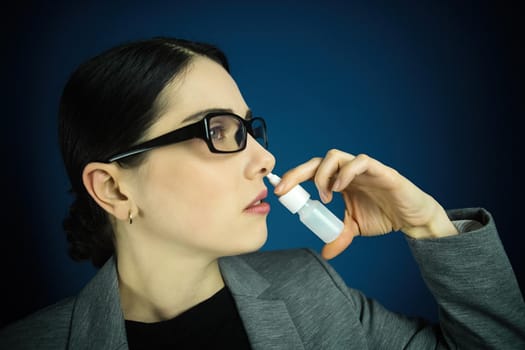 Young woman in glasses, using nose spray on blue background.