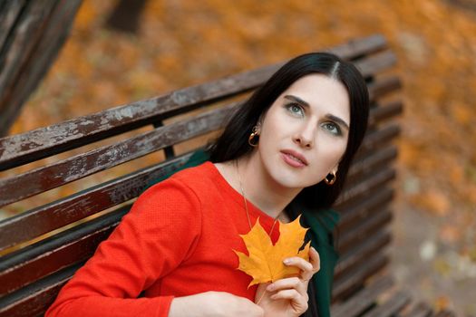 Young beautiful woman 25-35 years old sits on a bench in an autumn park with a large yellow leaf in her hands and looks into the sky..