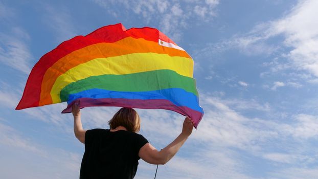 Bisexual, girl, lesbian, woman, transgender, homosexsual holding in hand a rainbow LGBTQIA gender identity flag on sky clouds background on a sunny day and celebrating a gay parade in pride month