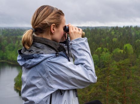 woman looks through binoculars. tourist girl on a rock in a forest near a lake