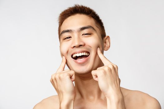 Beauty, skincare and men health concept. Headshot of happy, smiling enthusiastic asian man with naked torso, pointing fingers, poke cheeks and grinning, got rid of blemishes and acne.