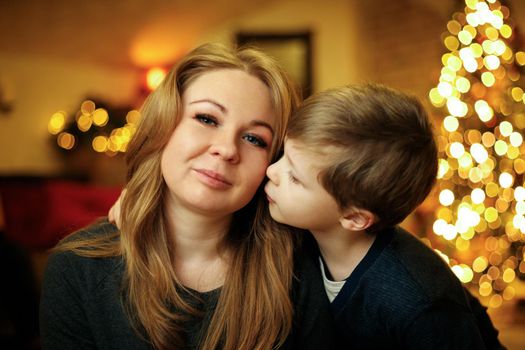 Boy 5-7 years old kisses his mother in a festive New Year's interior with a Christmas tree and garlands. Selective soft focus, film grain effect