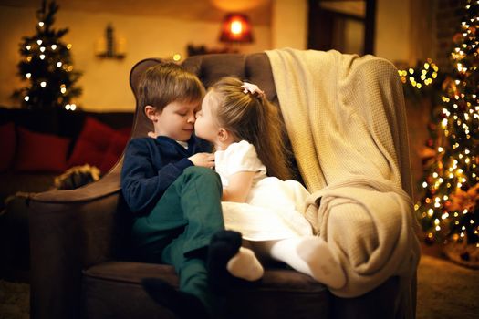 Little sister kisses his brother in a festive New Years interior with a Christmas tree and garlands. Selective soft focus, film grain effect