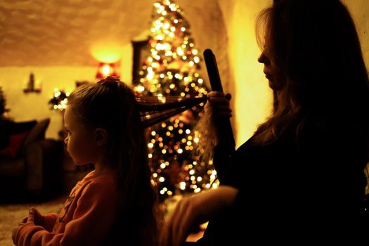 Mom combing her daughter for a festive dinner in the room with a christmas tree. Selective soft focus, film grain effect