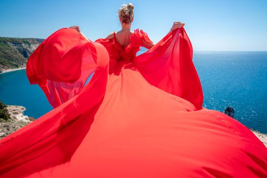 Blonde with long hair on a sunny seashore in a red flowing dress, back view, silk fabric waving in the wind. Against the backdrop of the blue sky and mountains on the seashore