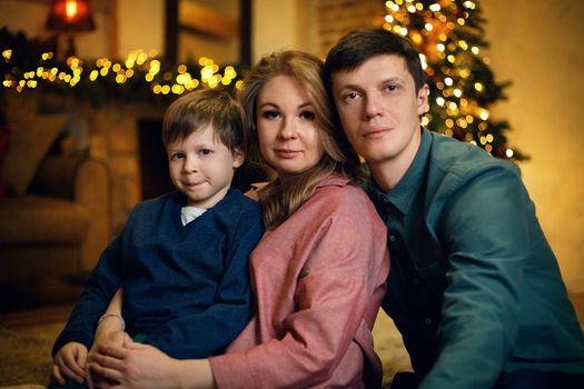 Portrait of young beautiful caucasian couple with their kid posing on floor in a cozy Christmas interior with a christmas tree and garlands. Selective soft focus, film grain effect